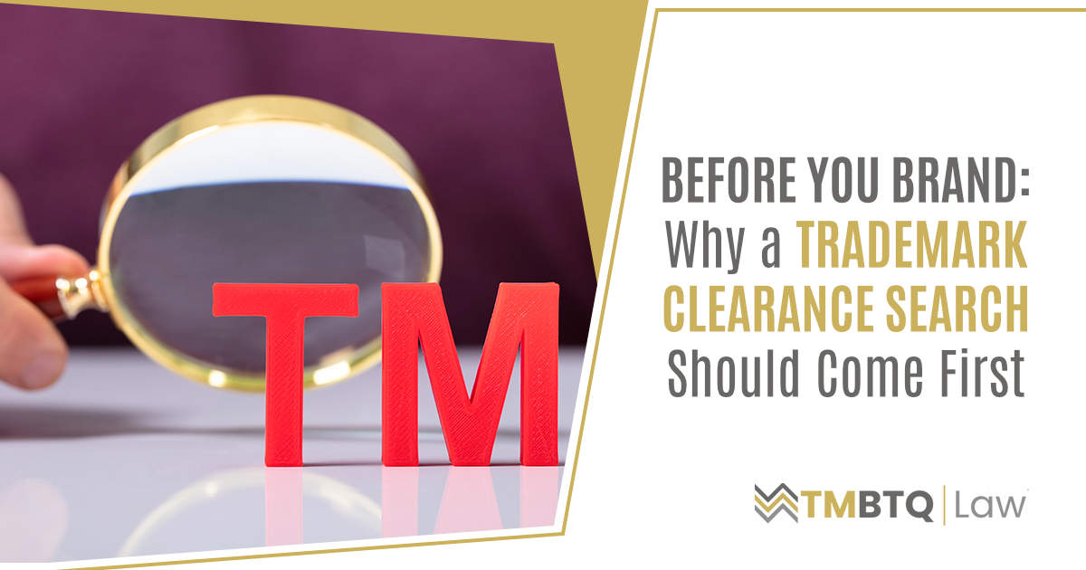 A hand holding a gold magnifying glass over large red 3D letters spelling 'TM,' symbolizing the trademark clearance search process before branding.