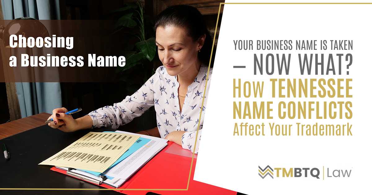 A professional woman in a patterned blouse reviews documents in an open binder at a wooden desk, representing the process of researching business name conflicts and trademark implications.